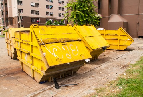 A row of large, yellow industrial rubble skips positioned on a paved area in an urban setting, with a backdrop of modern residential apartment buildings and a tree with lush green foliage. The skips are made of metal, featuring reinforced side walls, and show signs of dirt and minor wear. They are aligned side by side, with some positioned slightly in front of others, and are intended for waste collection or disposal. The concrete pavement beneath them has some small debris and grass patches nearby, indicating an outdoor environment possibly within a residential or commercial zone in Tooting or nearby South London. The overall scene reflects a typical waste removal setup managed by Tooting House Clearance for handling rubbish from property clearances or refurbishment projects.