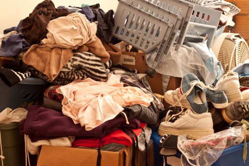 A large collection of mixed clothing items, shoes, and miscellaneous household textiles piled haphazardly on a flat surface, possibly for disposal or clearance. The textiles include various fabrics such as wool, cotton, and synthetic materials, with colours ranging from beige, cream, and black to red and maroon, some folded while others are crumpled. Several pairs of shoes are visible amidst the textiles, including sneakers and casual footwear, with some wrapped in plastic. Among the items are plastic storage baskets and cardboard boxes, partly filled with clothes and other soft items, with some stacked and leaning against the clutter. The background suggests an indoor space or an outdoor area with a wooden surface, typical of a domestic or clearance setting in Tooting or the surrounding South London area, where Tooting House Clearance operates. The scene conveys a sense of an ongoing clearance process, consistent with rubbish removal services that handle household waste and large textile collections.