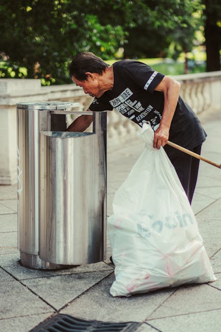An overflowing black metal trash bin with a domed lid is situated on a paved street, surrounded by various discarded items and litter. The top of the bin is cluttered with used paper cups, plastic cup