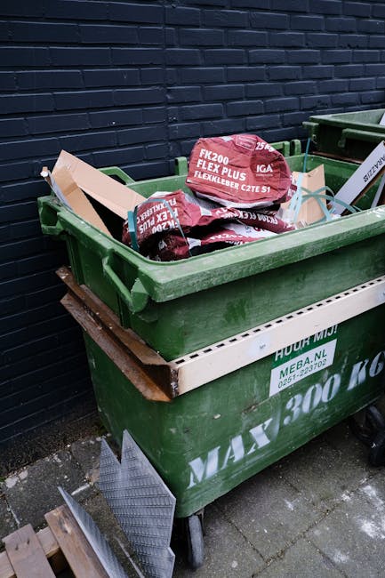 A woman with short dark hair, wearing a black T-shirt with white text on the front, is seen disposing of waste into a silver, metal, cylindrical rubbish bin situated on a paved sidewalk. She is holdin