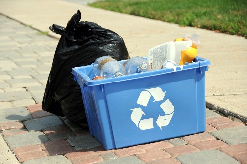 On a paved sidewalk near a grassy area, a blue rectangular recycling bin with a white recycling symbol is filled with clear plastic bottles, some with labels still attached. Next to the bin, there is a black refuse bag that appears to be full and tightly tied. The scene suggests a typical waste collection or rubbish removal situation, reflecting local rubbish disposal practices. The background shows a section of a curb and part of a green lawn, indicating an outdoor residential or public environment possibly in Tooting or nearby. The image is well-lit with natural daylight, emphasizing the different textures of the plastic bottles, the smooth surface of the recycling bin, and the slightly crumpled fabric of the refuse bag, aligning with the services offered by Tooting House Clearance for rubbish collection and waste disposal.